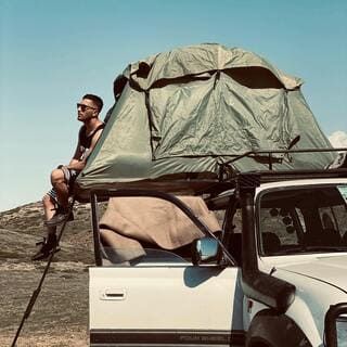 L'image montre un homme assis sur le toit d'un véhicule tout-terrain, qui est équipé d'une tente de toit. L'homme porte un t-shirt sans manches, un short et des lunettes de soleil. Il est assis avec ses jambes pendantes sur le côté du véhicule, regardant vers la gauche. Le véhicule est blanc et a une tente de toit verte déployée sur le dessus. Il y a un paysage de collines sèches et d'un ciel bleu clair en arrière-plan. L'ensemble donne une impression d'aventure et de plein air.