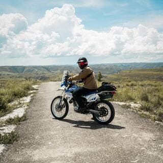 L'image montre une personne conduite une moto tout-terrain sur une route de gravier. La personne est vue de profil, portant un casque, une veste et des lunettes de soleil. La moto est blanche et bleue, avec un sac attaché à l'arrière. Le paysage environnant est vallonné, avec de l'herbe verte et quelques buissons. Le ciel est bleu avec de gros nuages blancs et cotonneux. La scène générale suggère une journée ensoleillée et une balade à moto dans la nature.