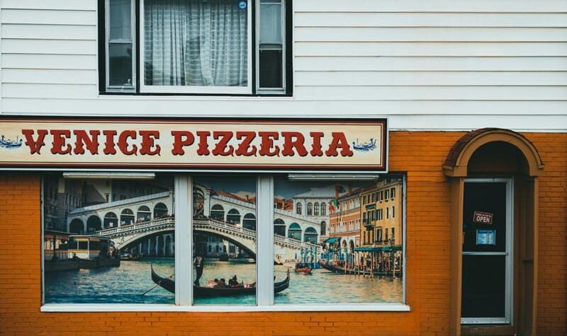 Une devanture de pizzeria nommée "Venice Pizzeria" avec une grande image ornant les fenêtres, représentant un canal vénitien avec des gondoles et des bâtiments de style italien. Au-dessus de la vitrine, une enseigne avec le nom de la pizzeria en lettres rouges stylisées, bordée de rouge et de bleu, sur un fond crème.