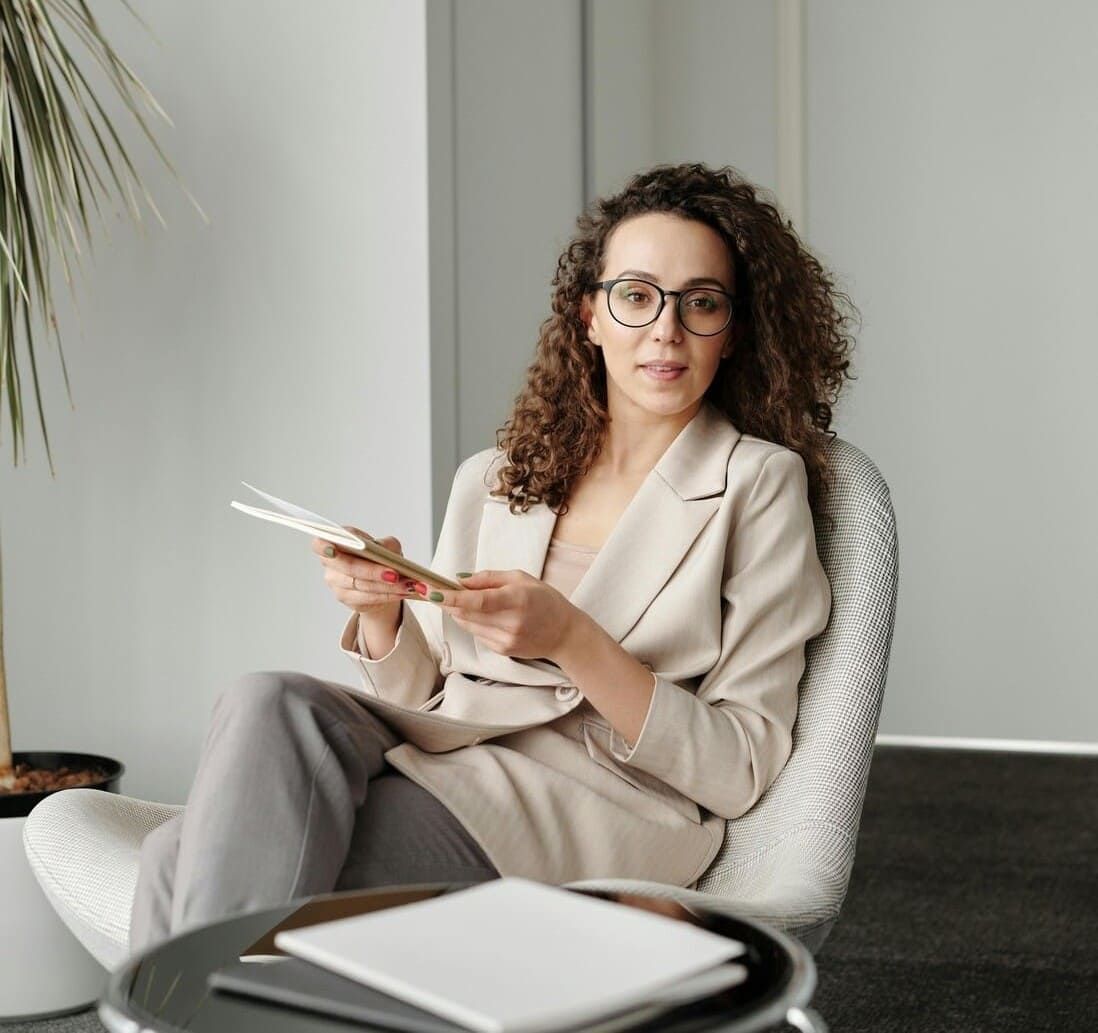 Une femme portant une veste beige, vous regarde avec le sourire, assise dans un fauteuil.