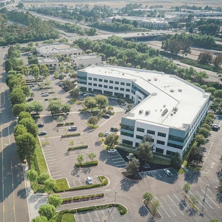 Un vaste parc de bureaux moderne avec un grand parking et des arbres luxuriants, en arrière-plan un bâtiment de bureaux à plusieurs étages.